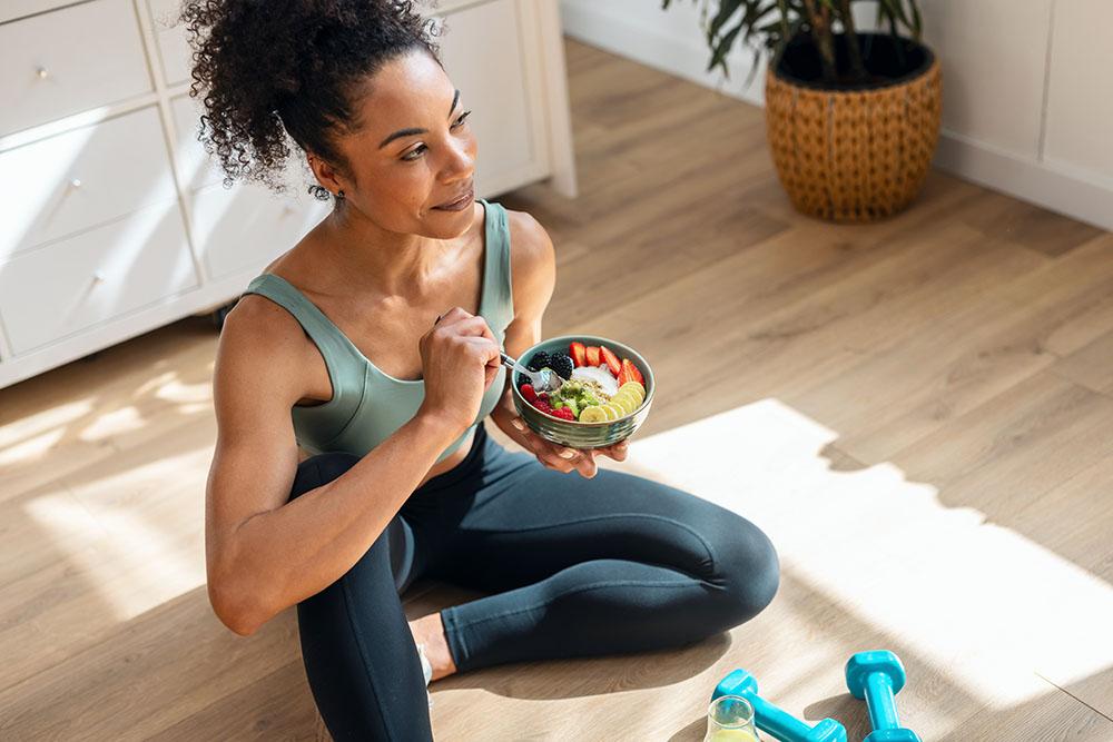 Woman eating yogurt with fruit on her living room floor in workout clothes
